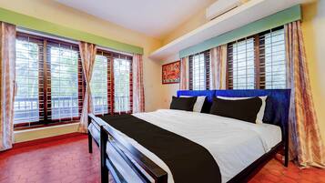 A hotel room with a bed, two windows with curtains, an air conditioning unit, and a painting on the wall.