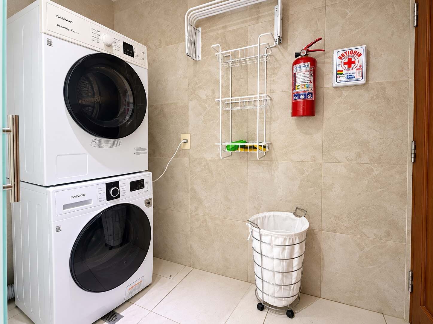 Een wasruimte met twee gestapelde wasmachines, een brandblusser, een verbanddoos en een metalen wasmand.