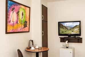 A hotel room with a painting, a television, a small table with a kettle, glasses, and a refrigerator.