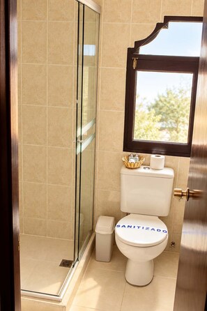 Een badkamer met een douchecabine van glas, een toilet met een 'SANITIZADO' label en een raam boven het toilet.