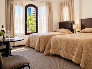 Ein Hotelzimmer mit zwei Betten, einem runden Tisch mit Flasche und Gläsern, einem Sessel und Blick auf Grünfläche.