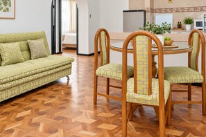 Un salon avec un canapé à motifs verts, des chaises en bois assorties à des coussins, une table ronde en verre et un parquet en bois disposé en chevrons.
