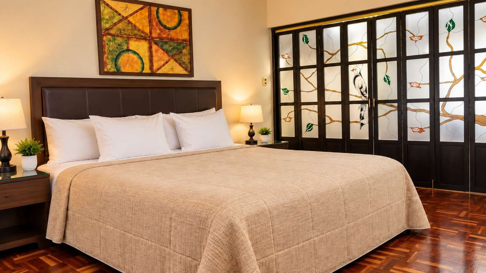 A neatly made bed with a beige bedspread, a dark headboard, and white pillows. There is a nightstand with a lamp and a small plant beside the bed.
