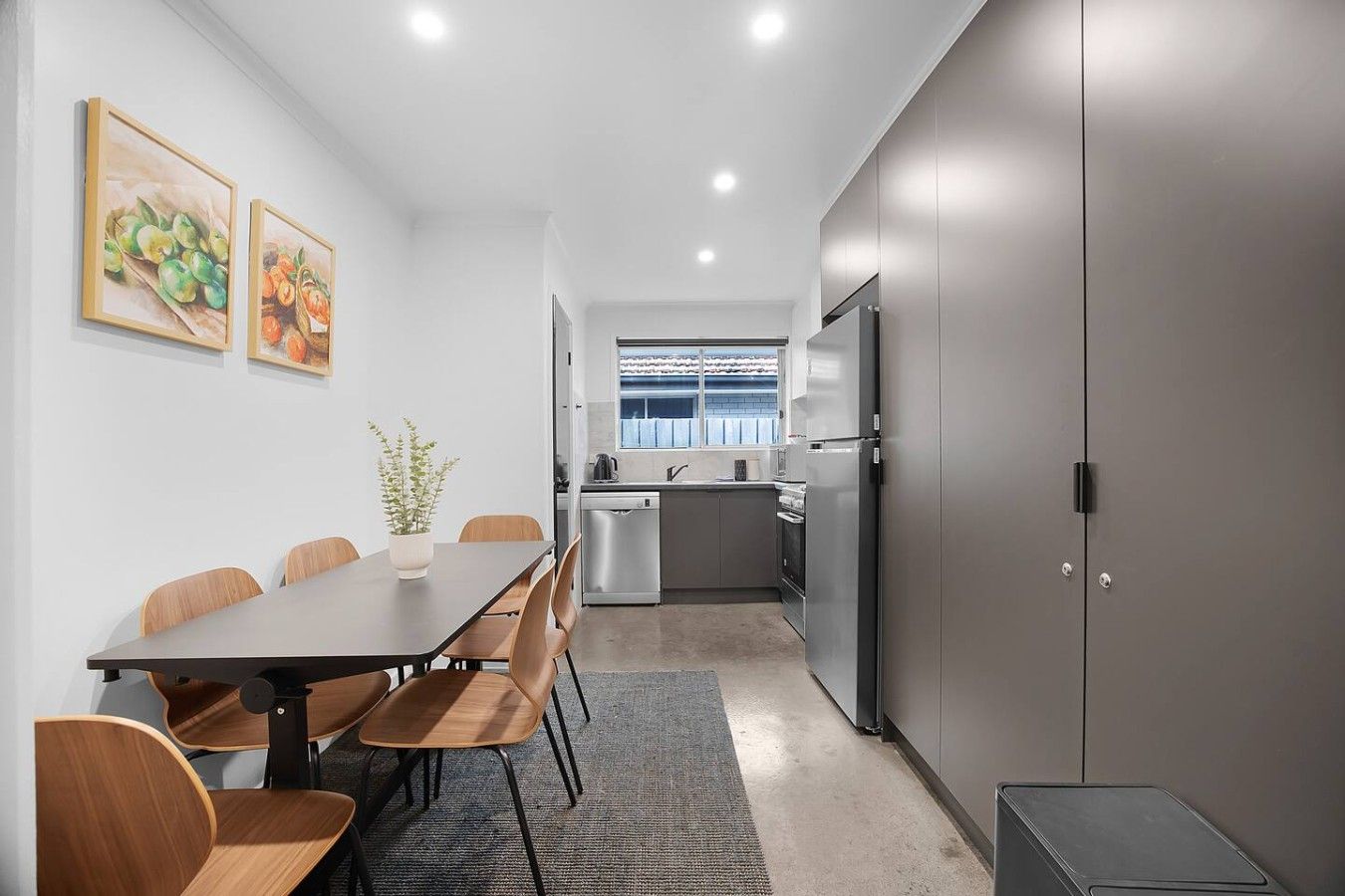 A modern kitchen with a dining area, featuring a long table with chairs, a refrigerator, and a window with a view.