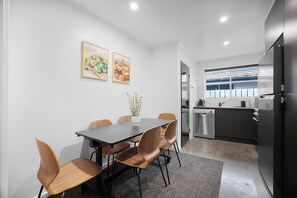 A modern kitchen with stainless steel appliances, a dining area with a table and chairs, and two framed artworks on the wall. - Duke’s Place in Sunshine (Sunshine North)