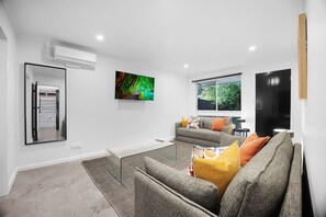 A modern living room with a glass coffee table, grey sofa with orange cushions, a painting on the wall, and a black door.