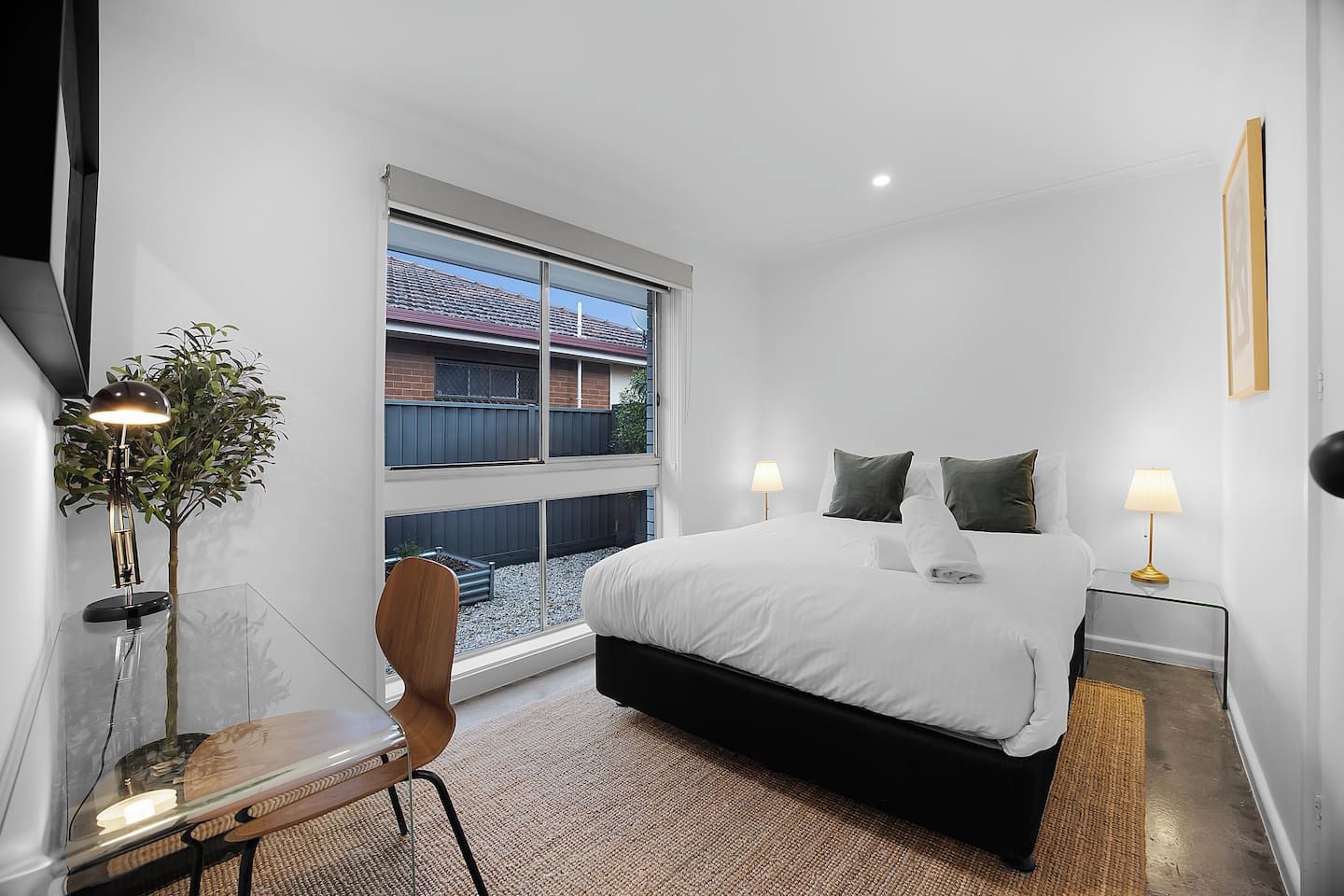 A modern hotel room with a large bed, bedside lamps, a desk with a chair, and a view of a residential area through the window.