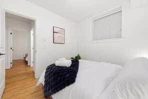 Un dormitorio con cama, ventana con persianas, puerta que da a otra habitación y un cuadro en la pared.