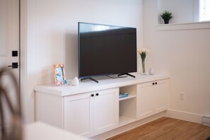 Una sala de estar moderna con un televisor de pantalla plana sobre un mueble blanco, una planta pequeña en el alféizar de la ventana y un jarrón con flores.