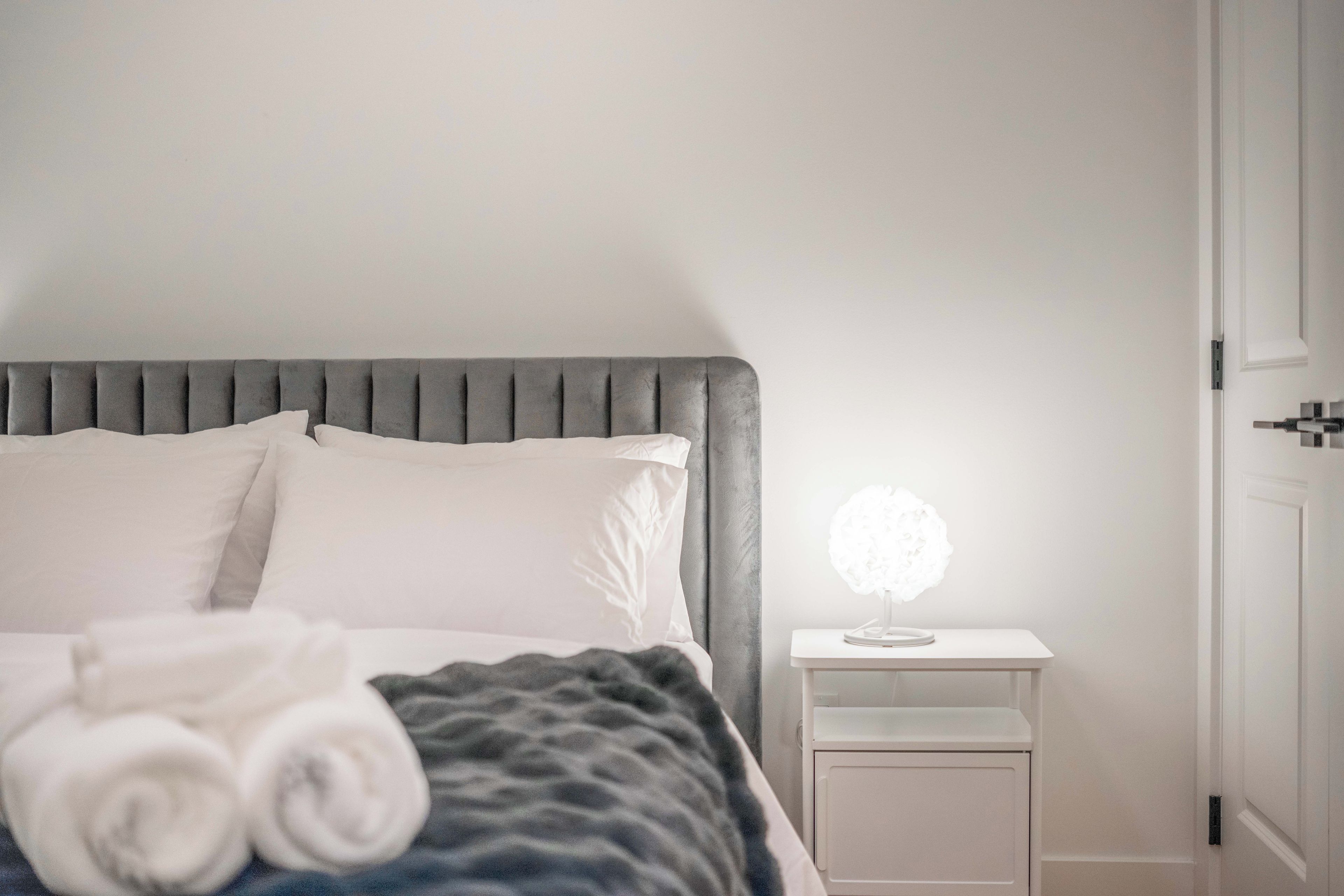 A neatly made bed with a grey headboard, a bedside table with a white lamp, and a door to the right.