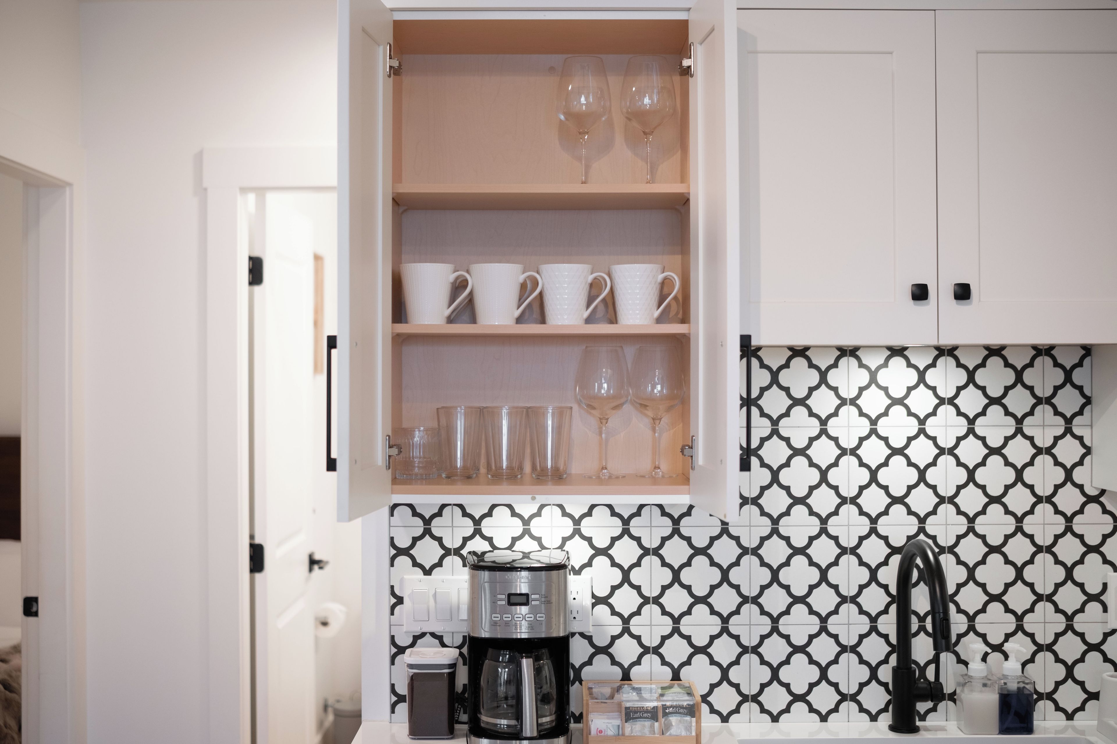 A kitchen with a coffee maker, a trash bin, and a cabinet with cups and glasses.