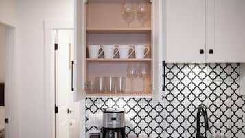 A kitchen with a coffee maker, a trash bin, and a cabinet with cups and glasses.