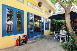A yellow building with blue-framed windows and doors, a covered outdoor seating area with white chairs and a table, and a fire extinguisher on the ground. - Loft House 2BR An Bang Beach Hoi An by Beachside Boutique Homes (Đà Nẵng)