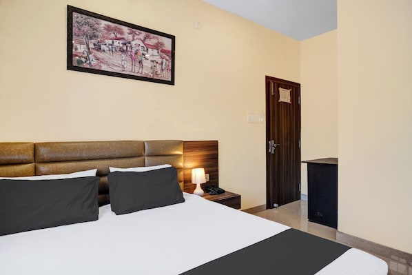 A hotel room with a large bed, a nightstand with a lamp, a wooden headboard, a door, and a painting on the wall. - Hotel O Lalganesh (Guwahati)