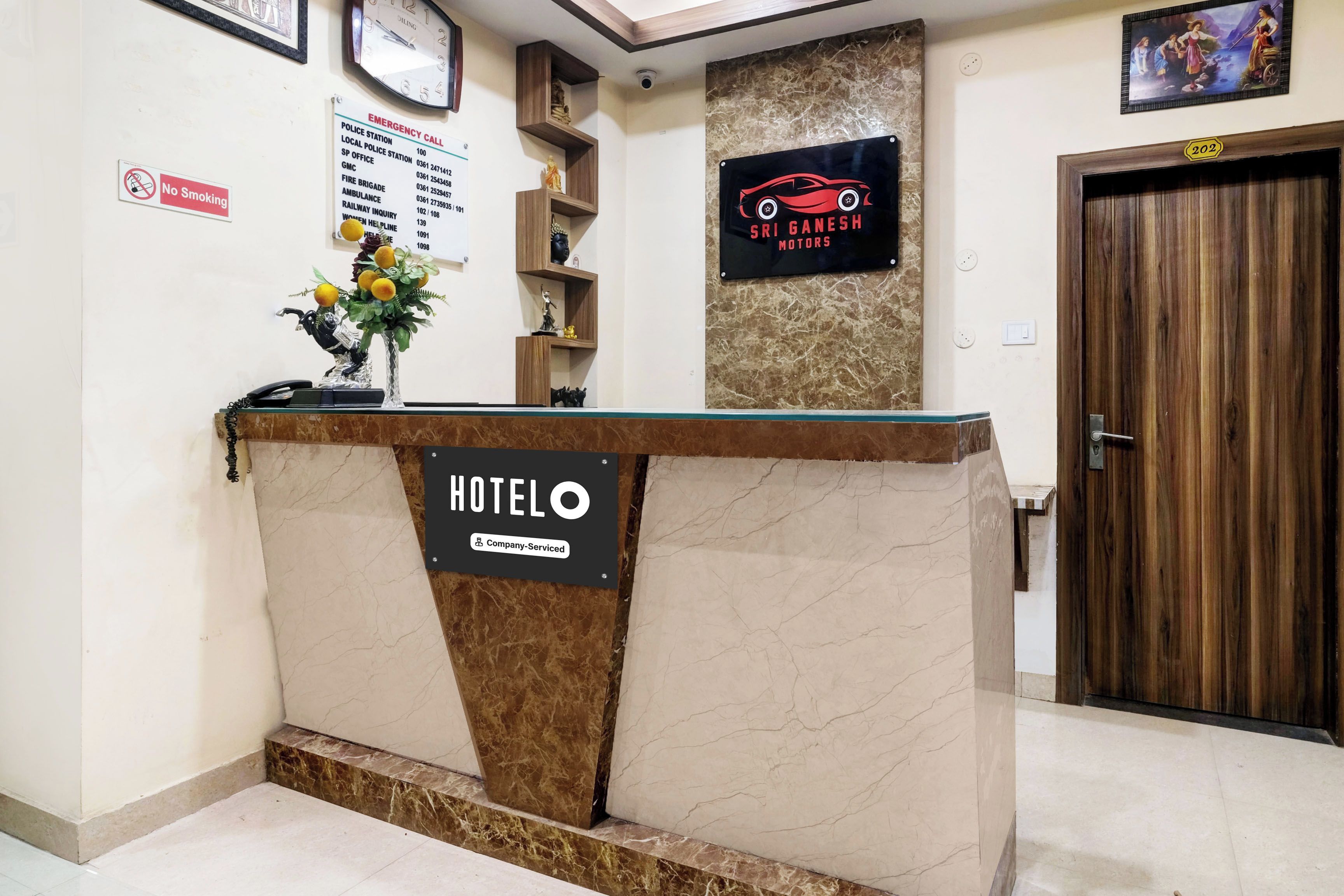 Una recepción de hotel con mostrador de mármol, un letrero que dice 'HOTEL', una etiqueta 'Servicio de Empresa' y un letrero de 'Sri Ganesh Motors'.