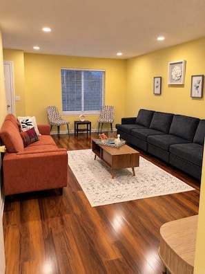 A living room with a red sofa, a dark grey sofa, a coffee table, two patterned chairs, a small side table, and a window with blinds. - Hidden Gem! Stylish 2BR Minutes from Dublin (Dublin)