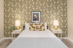 A bedroom with a floral wallpaper, a bed with white bedding, two bedside tables with lamps, and a framed picture above the bed. - Desert Hilltop Oasis in Foothills Epic (Tucson)