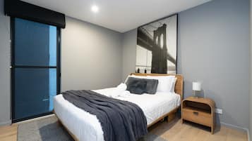 A modern bedroom with a large bed, a nightstand, a window with blinds, and a cityscape painting on the wall.