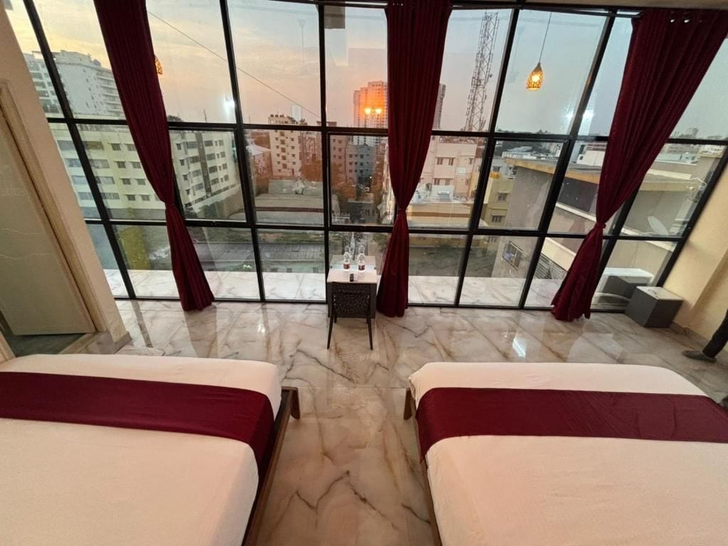 Ein Hotelzimmer mit zwei Betten, Blick auf die Stadt durch große Fenster und einem kleinen Tisch mit Stuhl.