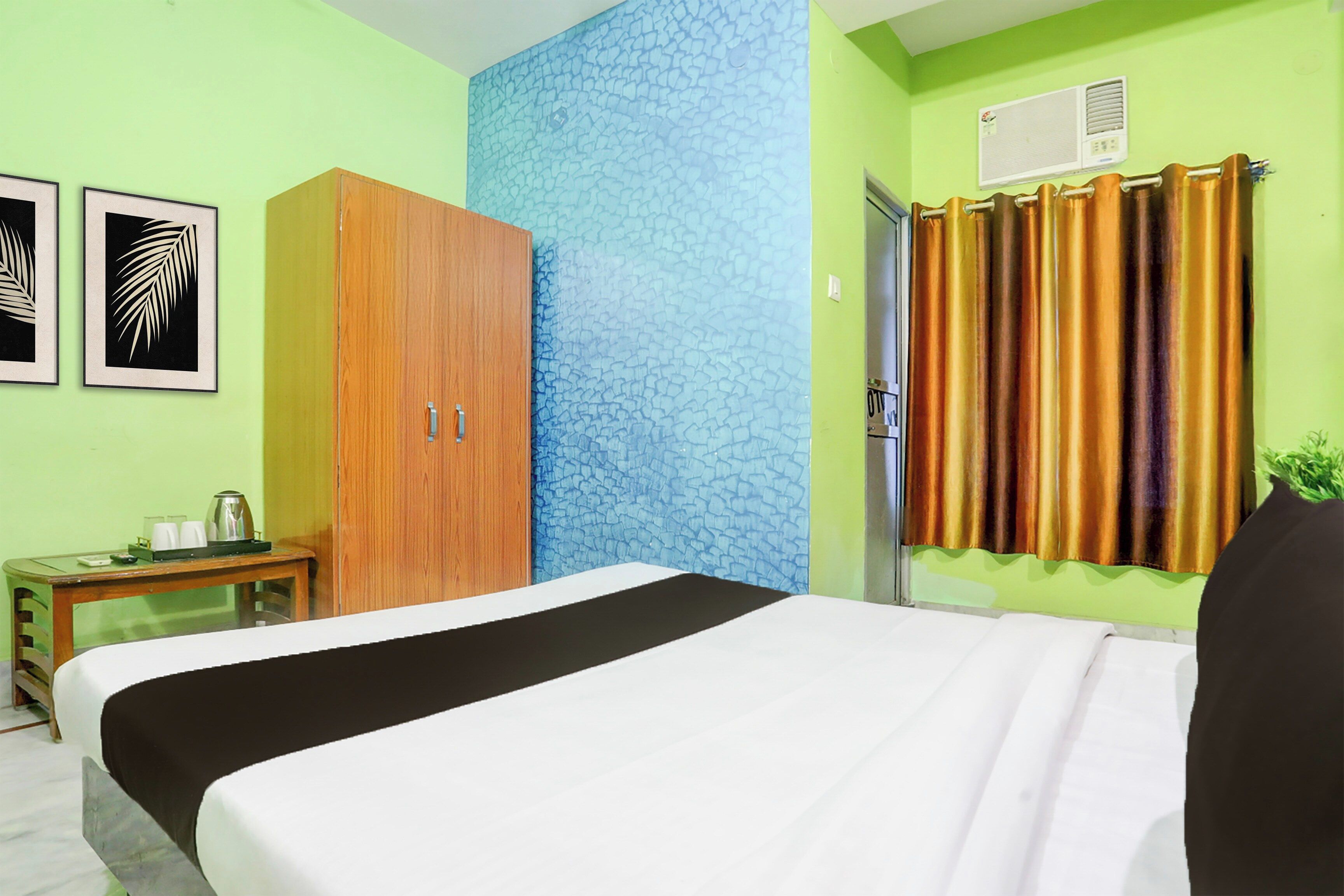 A hotel room with a bed, a wooden wardrobe, a small table with a kettle, and a window with curtains.