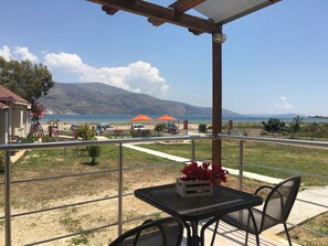 A balcony with a table and chairs, overlooking a beach with umbrellas and a mountain in the background. - Daphne's Apartment (Kefalonia)