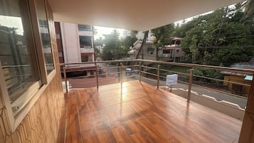 Un balcon doté d’un plancher en bois, d’une balustrade en verre et offrant une vue sur une rue bordée d’immeubles et d’arbres.