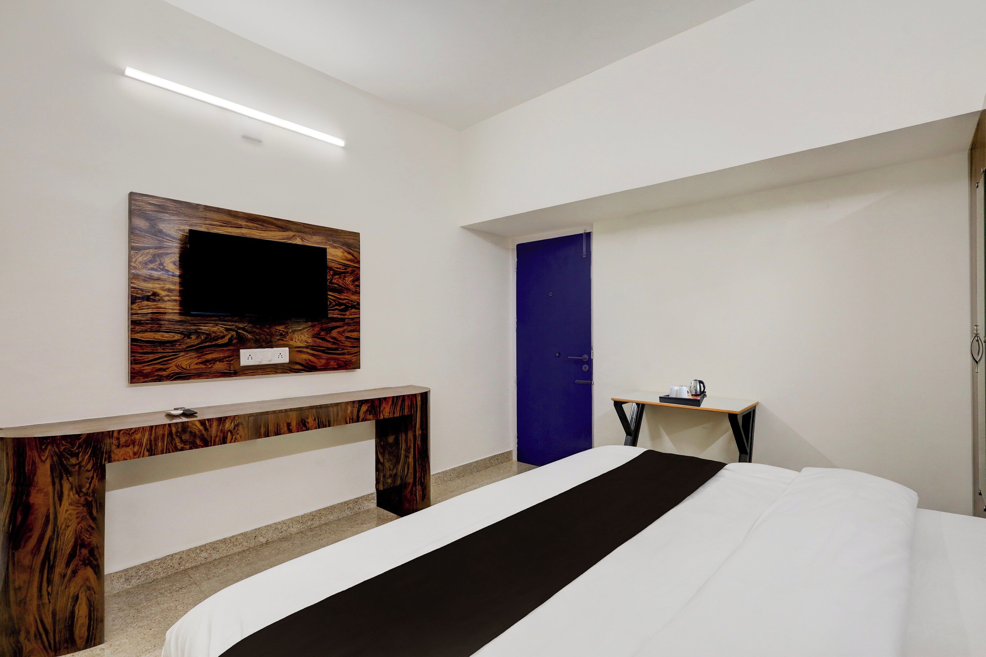 A hotel room with a bed, a TV mounted on a wooden panel, a small table with a tray, and a blue door.