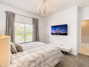 A bedroom with a large bed, a bench, a chandelier, a window with curtains, and a wall-mounted TV. - Golf Themed 4BR · Private Pool · ChampionsGate (Davenport)