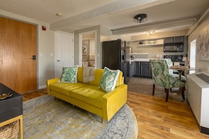 A yellow sofa with patterned cushions, a dining area with a table and chairs, a kitchen with appliances, and a wooden door. - Wall Street Suites 1 | King Suite near SIU with Kitchen (Carbondale)