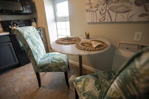 A kitchen with a dining area, featuring a chair with a leaf pattern, a round table with a woven placemat, and a wall-mounted artwork. - Wall Street Suites 1 | King Suite near SIU with Kitchen (Carbondale)