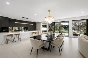 Cozinha moderna com ilha central, área de refeições com mesa e cadeiras de madeira escura, e janelas amplas com vista para o exterior.