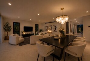 A modern living room with a dark wooden dining table, beige sofas, and a large flat-screen TV. - Riverfront Retreat : Pool, Cinema & Putting Green (Benowa)
