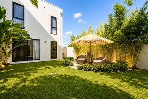 A modern house with a well-maintained lawn, outdoor seating with a striped umbrella, and bamboo plants. - Riverfront Retreat : Pool, Cinema & Putting Green (Benowa)