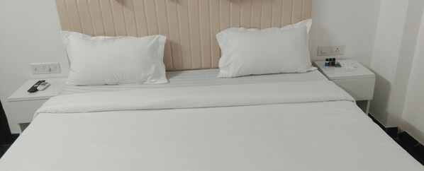 Habitación de hotel con una cama, dos almohadas blancas, un control remoto y una mesita de noche con una botella y una caja.