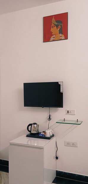 Una habitación con una televisión de pantalla plana montada en la pared, un mueble blanco pequeño con una tetera y una cafetera, y un estante de vidrio con un control remoto.