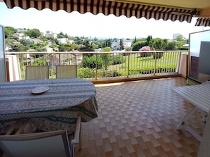 A balcony with a tiled floor, a table with a patterned tablecloth, and a view of a green landscape with buildings in the distance. - Holiday Apartment With 2 Bedrooms (Antibes)