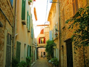 A narrow street with buildings on either side, a visible shop at the end, and a small table with chairs outside. - Holiday Apartment With 2 Bedrooms (Antibes)