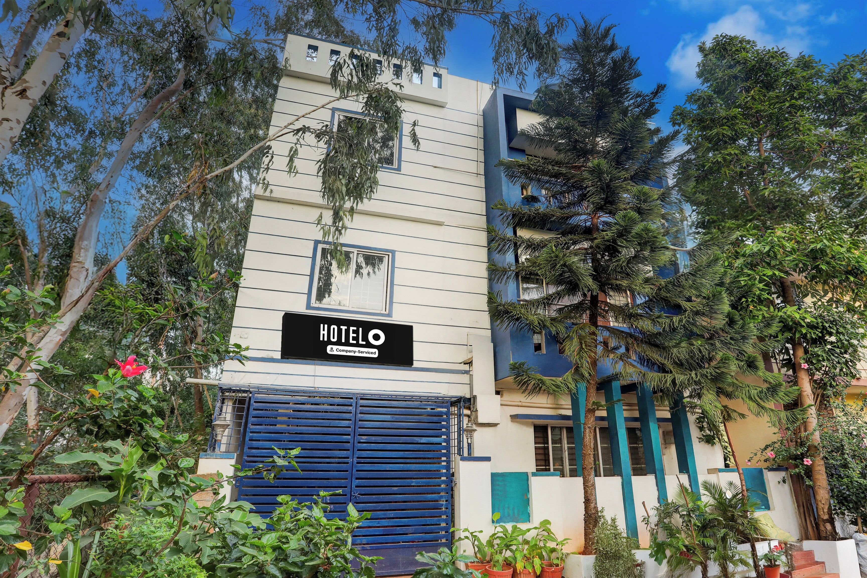 Un hotel con un letrero que indica que ofrece servicios para empresas, una verja azul y varias plantas y árboles alrededor del edificio.