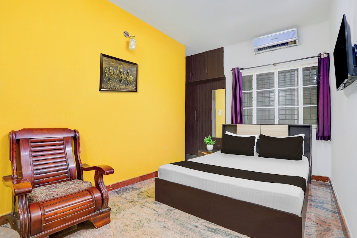 Una habitación de hotel con cama, una silla de madera, una ventana con cortinas, un equipo de aire acondicionado y un televisor montado en la pared.