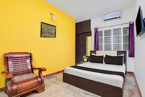 Una habitación de hotel con cama, una silla de madera, una ventana con cortinas, un equipo de aire acondicionado y un televisor montado en la pared.