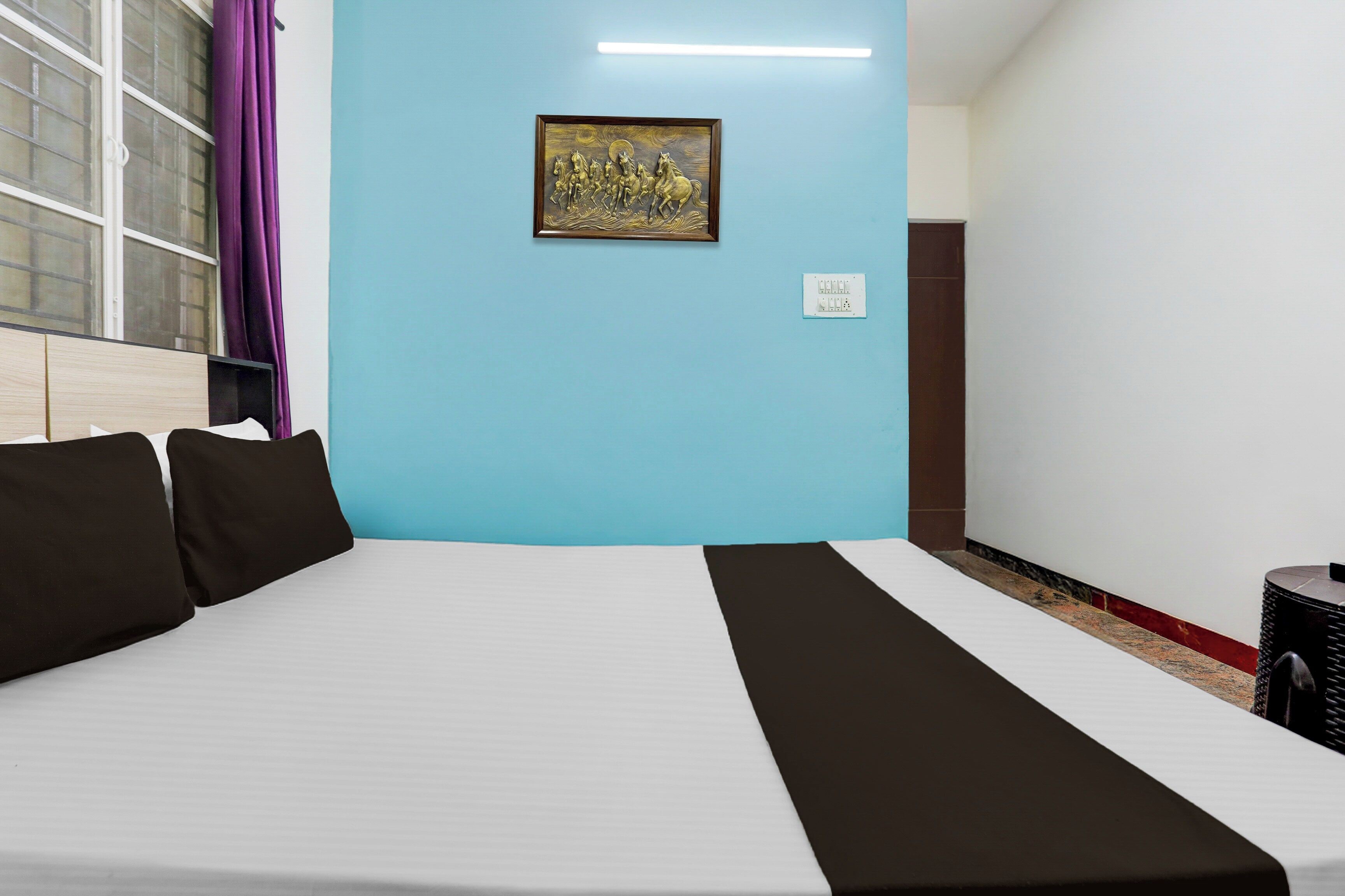 Una habitación de hotel con una cama, una lámpara de pared, un enchufe y una ventana con cortinas.