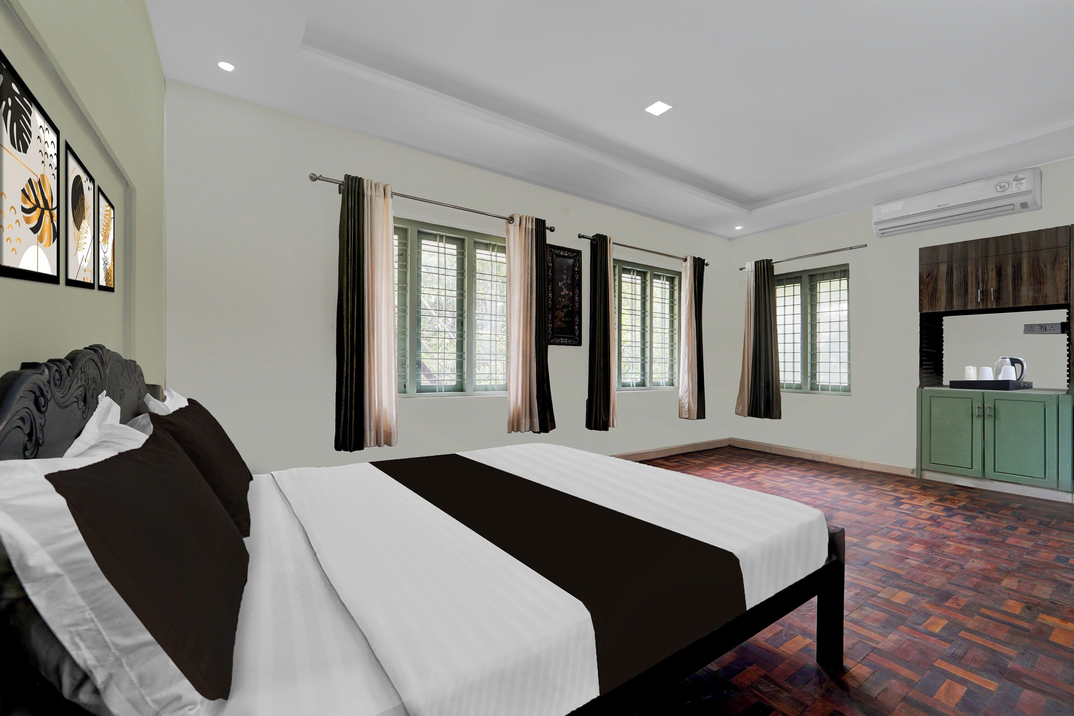 Quarto de hotel com uma cama, duas janelas com cortinas, uma kitchenette e obras de arte na parede.