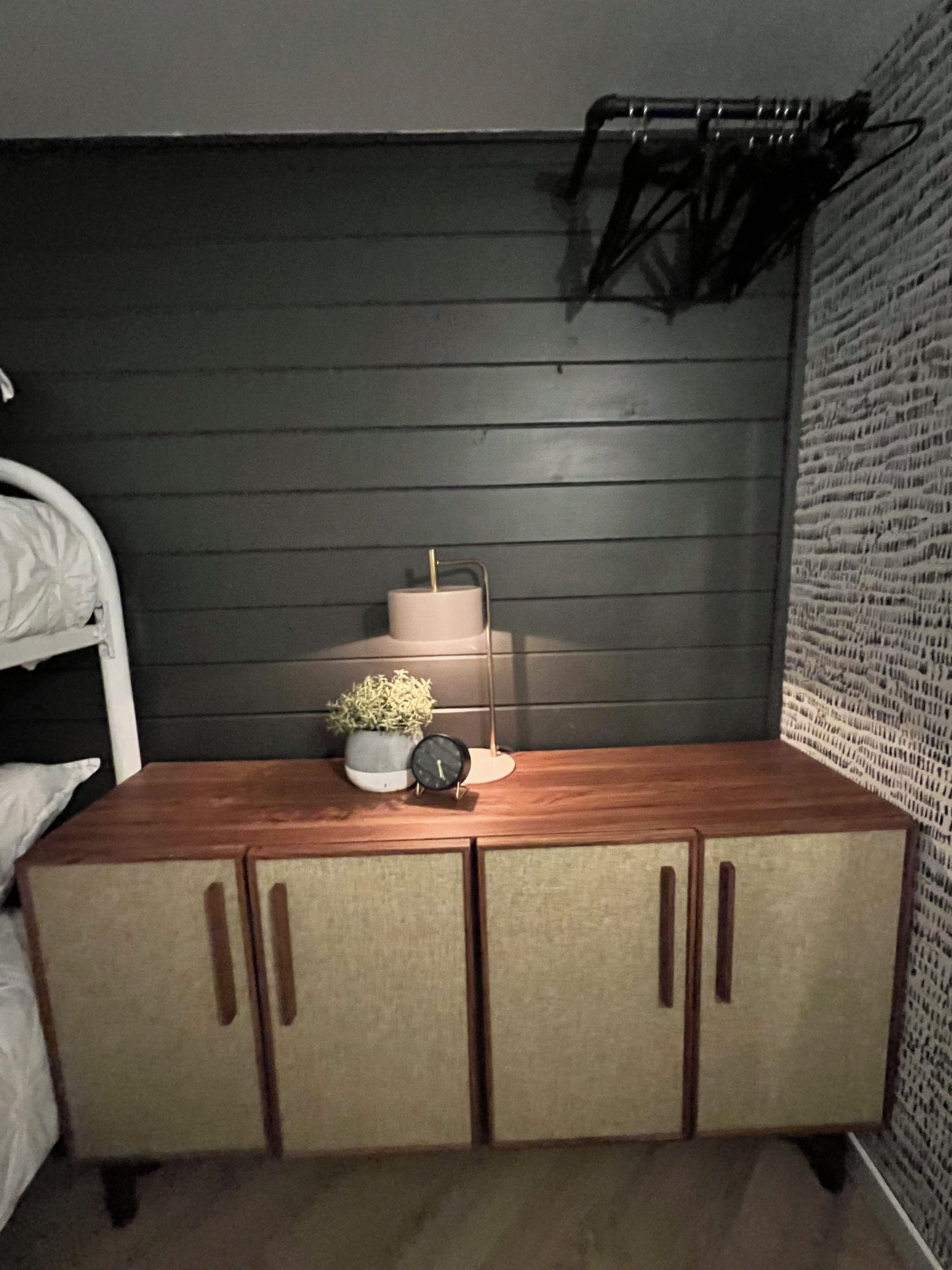 A bedroom with a wooden bed, a bedside table with a clock, a small plant, and a wall-mounted rack.