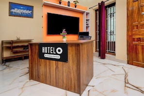 Una recepción de hotel con un cartel que indica servicio de empresa. Hay un televisor de pantalla plana montado sobre el mostrador, una planta en maceta y una puerta de madera a la derecha.