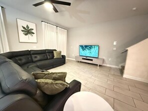 A living room with a black leather sofa, a white coffee table, a flat-screen TV on a stand, and a ceiling fan. - LUXURY 5BR PRIVATE POOL 8 MIN TO DISNEY SLEEPS 13 (Kissimmee)