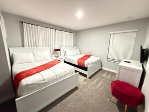 A hotel room with two beds, a small table, a red chair, a TV, and a window with blinds. - LUXURY 5BR PRIVATE POOL 8 MIN TO DISNEY SLEEPS 13 (Kissimmee)