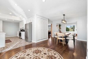 A modern kitchen with white cabinetry, a stainless steel refrigerator, and a dining area with a wooden table and chairs. - Hillside Family Retreat • 3BR/2BA • Near the Zoo (Oakland)