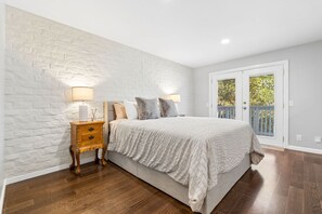 A bedroom with a white brick wall, a wooden nightstand with a lamp, a bed with a white duvet, and a view of a balcony with a blue railing. - Hillside Family Retreat · 3BR + Library · Near the Oakland Zoo (Oakland)
