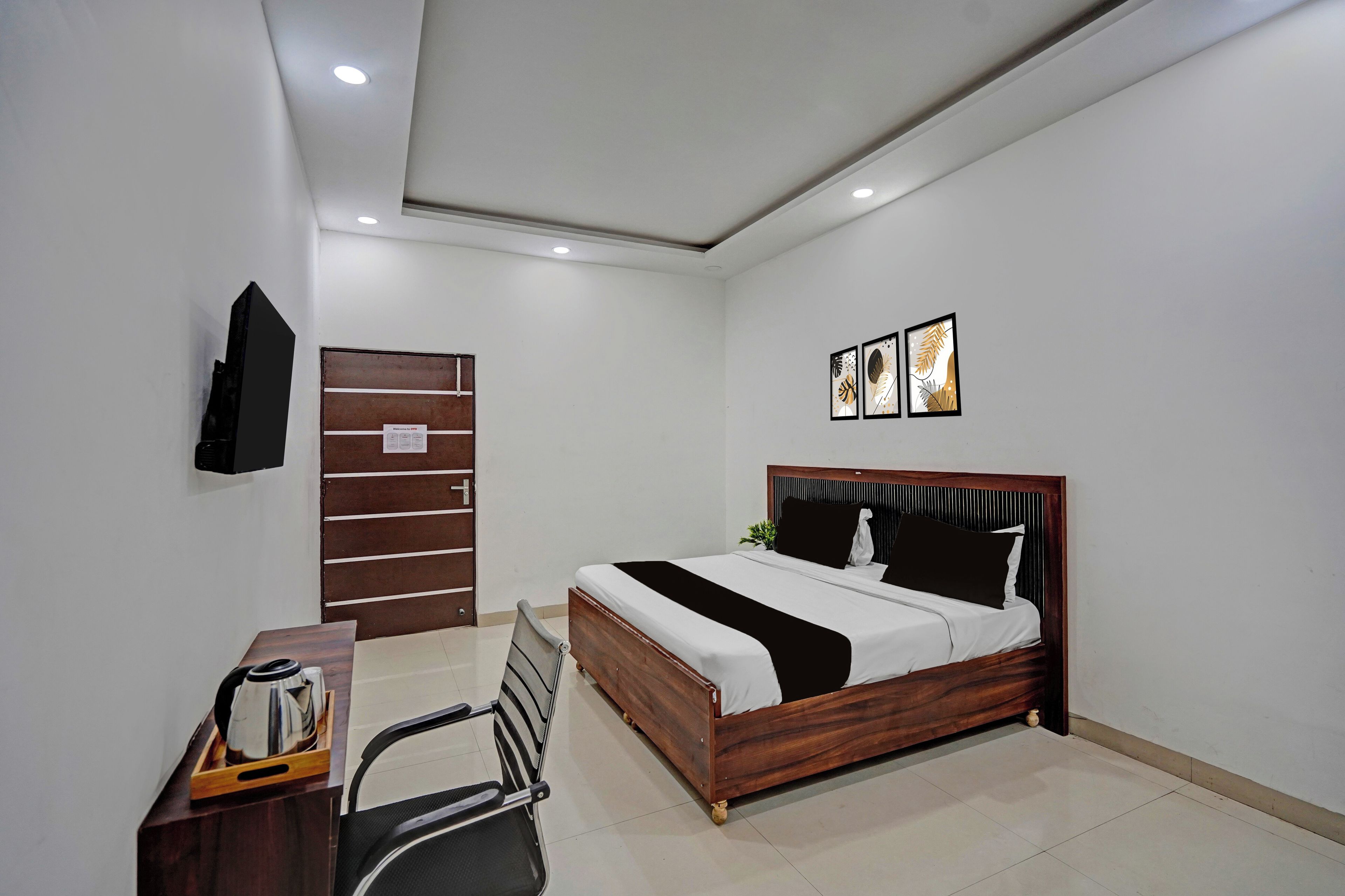 Un dormitorio con cama de madera, un escritorio con hervidor, una silla y un televisor en la pared.