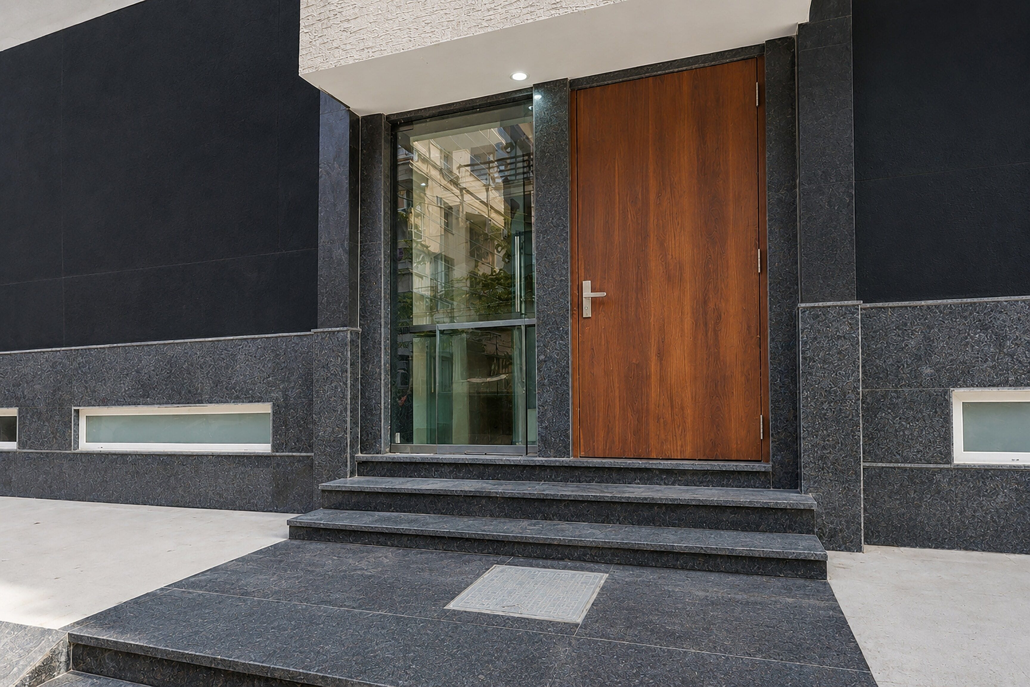 A modern building entrance with a glass door and a wooden panel, flanked by dark stone walls and small windows.